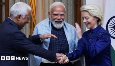 Three leaders are standing close together indoors, smiling and laughing during a friendly moment. The man in the centre, Indian Prime Minister Narendra Modi, with a white beard and glasses, wears a light shawl over a dark outfit and appears animated. On either side, European Council President António Luís Santos da Costa (left) in a dark suit and European Commission President Ursula von der Leyen (right) a deep blue jacket lean in, clasping hands in a gesture of warmth. A national flag and curtains are visible behind them