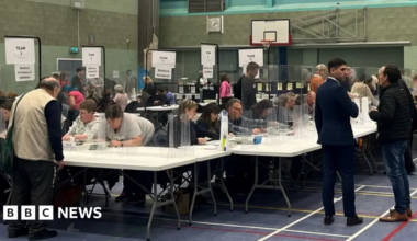 Counters go through ballots at a previous Cherwell District Council election count. They sit at white trellis tables in a sports hall, behind plastic screens. Candidates and members of political parties watch the count.
