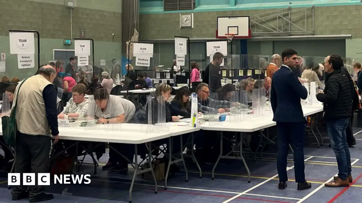 Counters go through ballots at a previous Cherwell District Council election count. They sit at white trellis tables in a sports hall, behind plastic screens. Candidates and members of political parties watch the count.