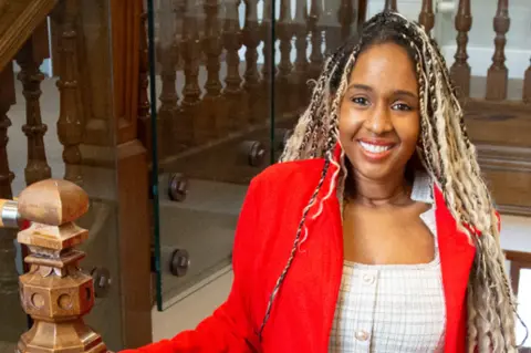 Kiki Marriott is stood inside the Parkland Place Rehabilitation Centre at the top of some wooden stairs. She has blonde braids and is wearing a white patterned suit with a red jacket. Kiki looks directly at the camera.