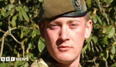 A man with blonde hair and a greenish brown beret with bushes behind him