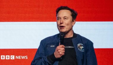 A woman wearing a sleeveless, yellow textured top, seated in front of a mirror with round vanity lights in a dressing room setting.