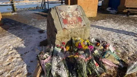 BBC/Becki Bowden A close up of flowers laid next to a memorial stone near the beach.