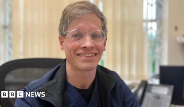Dr Chris Jacobs smiles at the camera. He is wearing glasses, a blue gilet and medical scrubs top. He sits in a computer chair.