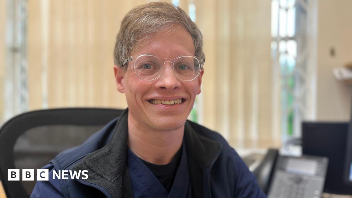 Dr Chris Jacobs smiles at the camera. He is wearing glasses, a blue gilet and medical scrubs top. He sits in a computer chair.