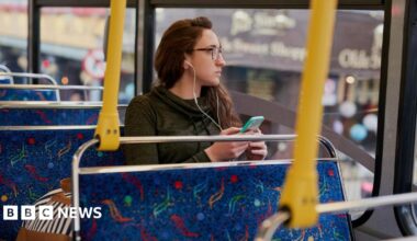 A young woman wearing glasses looks out the window of a bus while listening to her phone using headphones.
