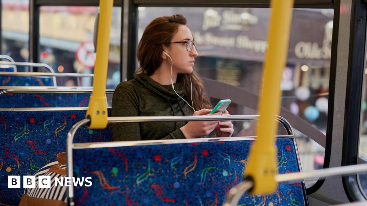 A young woman wearing glasses looks out the window of a bus while listening to her phone using headphones.