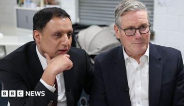 Anas Sarwar, in a dark blue suit and white shirt with a burgundy tie, sits with his chin resting on his hand. To his left, on the right of the shot, is Sir Keir Starmer in a blue suit and white open-necked shirt. Sir Keir is wearing thin, black plastic glasses. They are looking in the same direction off screen and appear to be listening. They are in a white, functional room with a pram behind them.