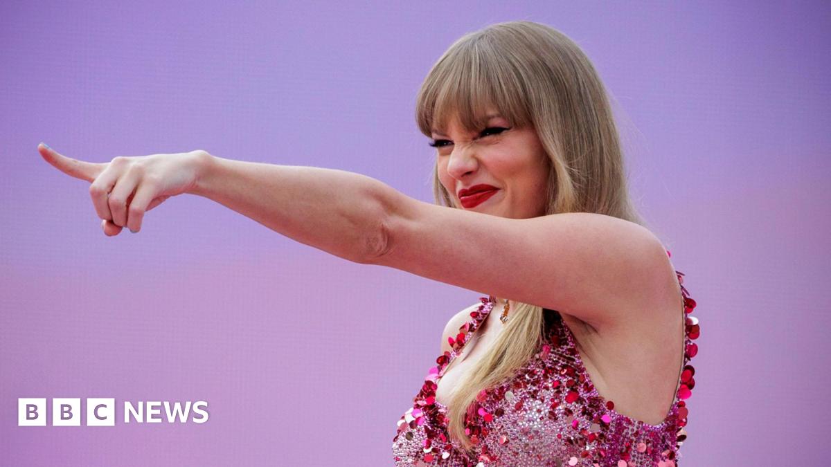 Taylor Swift points into the distance with a grin and her eyes squinted beneath her blonde fringe. She wears a sparkly pink fitted bodice and red lipstick. The background is pinkish-purple gradient and matches her outfit.