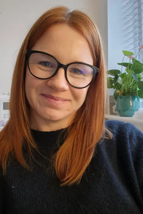 Hayley Rawlings Hayley Rawlings smiles at the camera. She has shoulder-length red hair and wears a blue knitted jumper. She has black-framed glasses on. It is a selfie, and a head and shoulders shot.