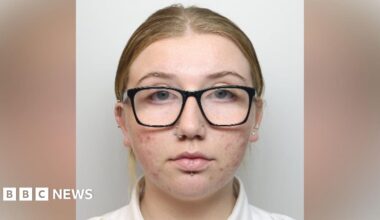 A woman with black-framed glasses, two nose piercings and a lip piercing looks at the camera.