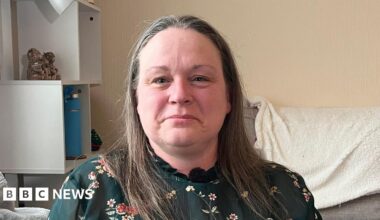 Elaine is looking at the camera. She has long brown/grey hair and is wearing a green floral top with a high neckline.