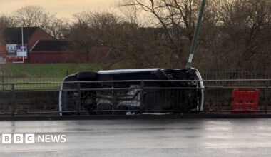 A car on its side, with its underside towards the camera, behind a pedestrian barrier next to a road, with parkland visible beyond