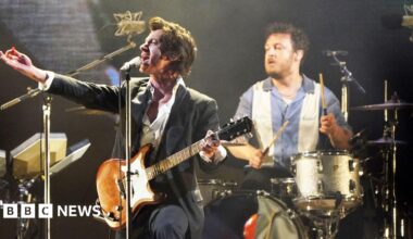 Frontman of Arctic Monkeys Alex Turner plays guitar on stage and drummer Matt Helders is seen closely behind him playing drums. Matt Helders is wearing a blue and white striped shirt and Alex Turner is wearing a black suit jacket and white shirt