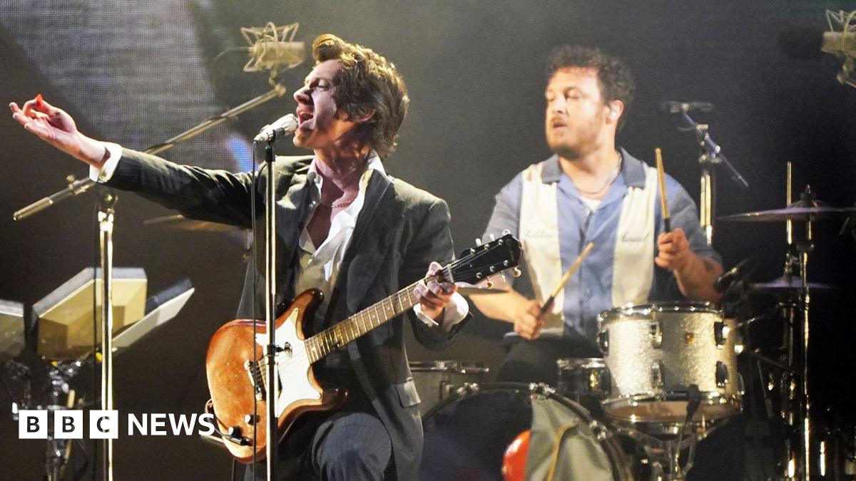Frontman of Arctic Monkeys Alex Turner plays guitar on stage and drummer Matt Helders is seen closely behind him playing drums. Matt Helders is wearing a blue and white striped shirt and Alex Turner is wearing a black suit jacket and white shirt