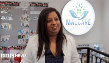 A woman in a jacket looking to the side of the image. Behind her is a display board with lots of small photographs and a large, lit up logo that reads Maucare.