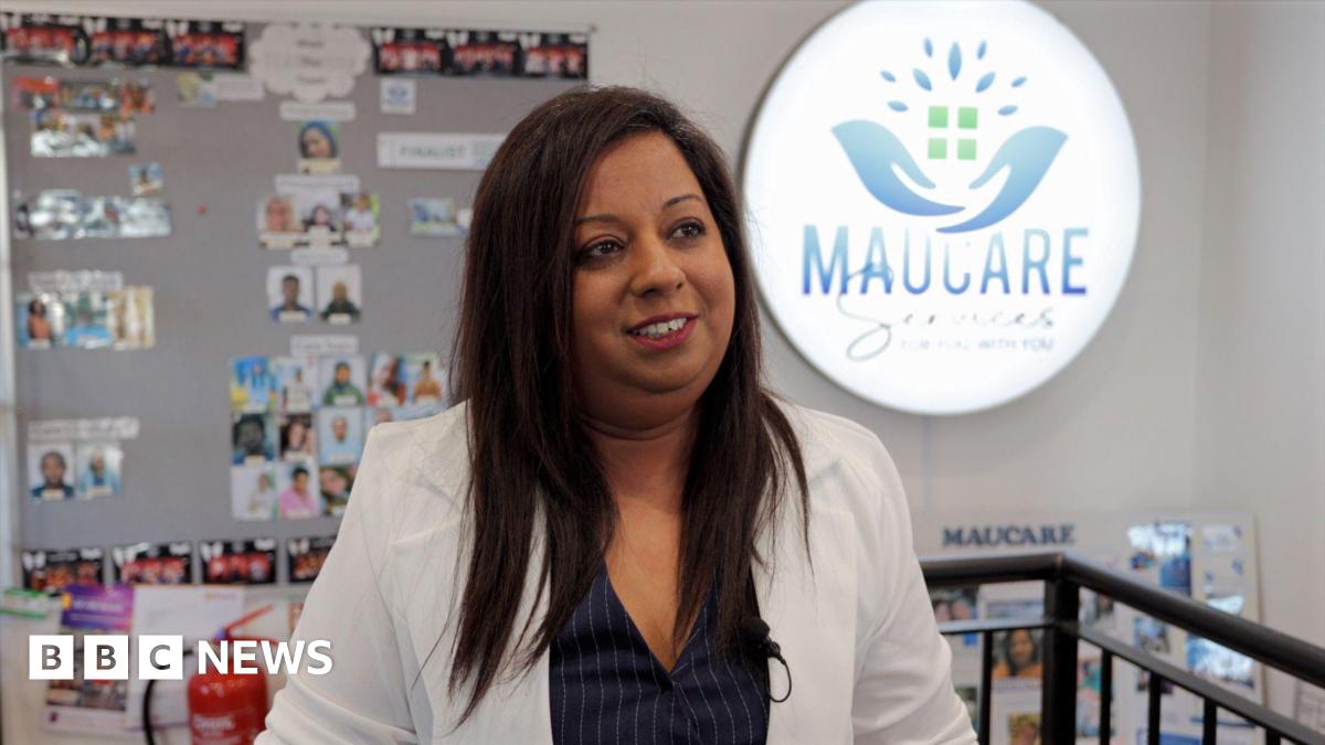 A woman in a jacket looking to the side of the image. Behind her is a display board with lots of small photographs and a large, lit up logo that reads Maucare.