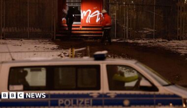 Police and emergency personnel wearing uniforms and one a helmet inspect a metal box with graffiti on it beside a fence in the dark. A police van is seen in the foreground and snow is on the ground.