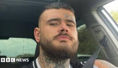 A selfie of a man sitting in a car. The trees in the background are blurred showing the car is moving. He is wearing a seatbelt and looking into the lens. He has a bear, a neck tattoo and a small tattoo above his eyebrow.