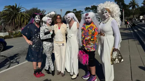 California Academy of Sciences Press Office Five drag queens, most dressed in white, standing and smiling with a woman 