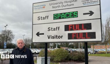 Parish councillor Grenville Holland, wearing a dark winter coat, is standing in front of a sign for the car park. A full car park is visible in the background. The sign itself if white and has digital elements. The digital parts display the words "full" in red next to an arrow to visitor and staff parking. An arrow pointing in the opposite direction labelled 'staff' says 'spaces' in green.