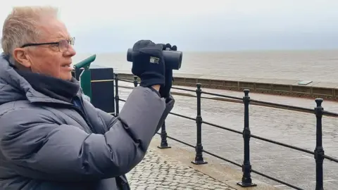 Stuart Baines A man with grey hair and black rimmed spectacles holds up a set of binoculars to his face. He is wearing a grey  puffa jacket and standing on a cobbled pavement behind black metal railings looking out to sea. In the background is a road a low wall and a grey sea stretching to the horizon and a light blue sky.