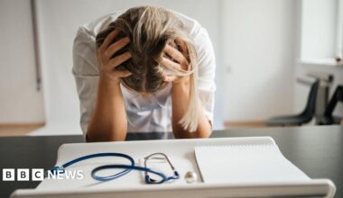 A stock image shows a female doctor with long blonde hair leaning on a black desk with her head in her hands so only the top of her head is visible - not her face. She is wearing a white dress or lab coat. In the foreground is a blue stethoscope and a white paper writing pad. The room has white walls and a black chair can be seen the back right corner of the image.
