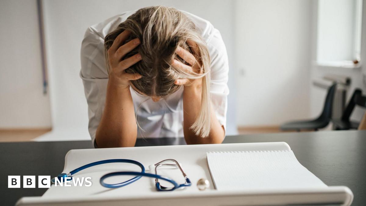 A stock image shows a female doctor with long blonde hair leaning on a black desk with her head in her hands so only the top of her head is visible - not her face. She is wearing a white dress or lab coat. In the foreground is a blue stethoscope and a white paper writing pad. The room has white walls and a black chair can be seen the back right corner of the image.