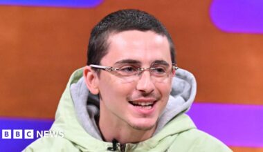 Timothée Chalamet, who has cropped dark hair and wears glasses, smiles as he appears on sofa of the Graham Norton chat show. He is front of a orange and purple backdrop.