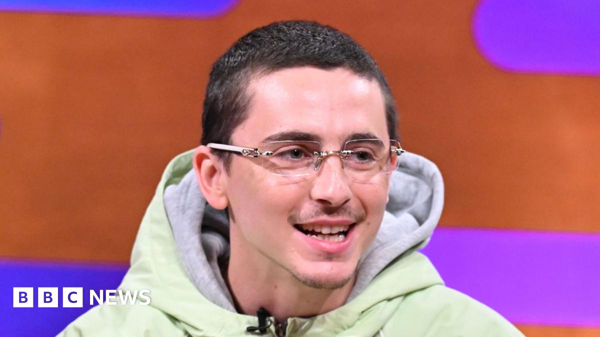 Timothée Chalamet, who has cropped dark hair and wears glasses, smiles as he appears on sofa of the Graham Norton chat show. He is front of a orange and purple backdrop.