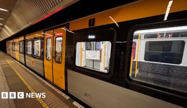 A yellow, grey and black Metro train pulled into a station. The train is empty and its destination is Pelaw which is lit up on an LED display by the doors. The platform has a brown and yellow tiled floor.