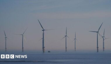 Millie Bobby Brown and David Harbour in character as Eleven and Jim Hopper. Eleven wears red shorts over grey leggings and a grey jumper. Jim Hopper wears a black leather jacket, a black beanie hat and dark trousers.They are speaking to director Frank Darabont on the set of Stranger Things. There is a vintage advertisement posted in the background with debrief on the ground.