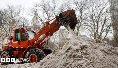 An orange plow dumps snow onto a large, grey snow pile in New York City.  City Hall can be seen in the background.