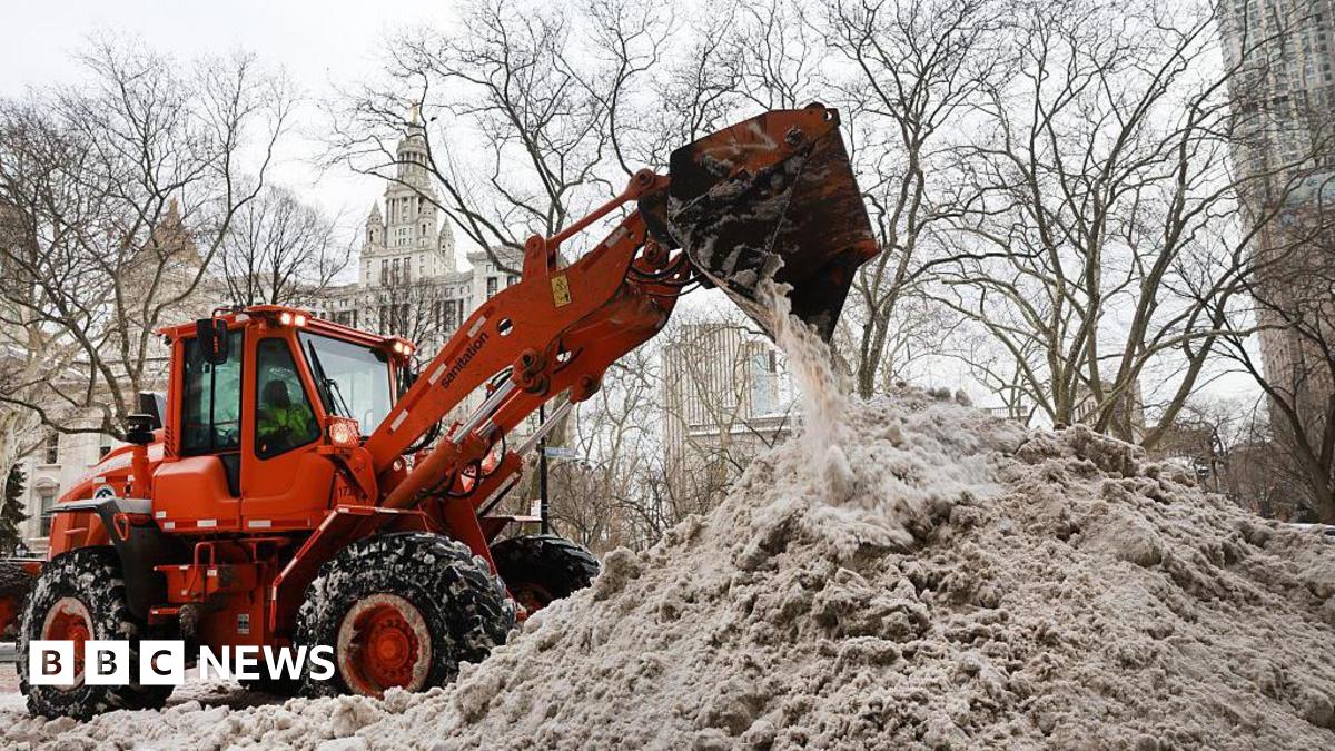 An orange plow dumps snow onto a large, grey snow pile in New York City.  City Hall can be seen in the background.