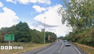 A road with vehicles travelling in both directions. There are street lights and green road signs at the side of the road as well as a pavement running for some of its length.