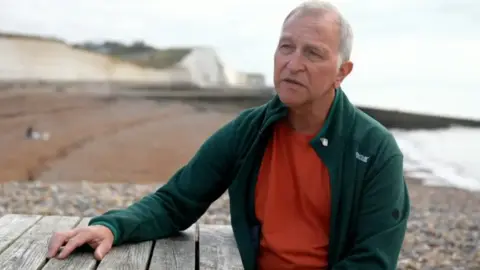 Giles Turner Photo of Giles Turner who has white hair and is wearing a red top and green fleece. His hand is resting hand on a table. There are cliffs, beach and sea in the background. 