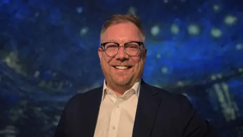 Ben Ward, a man wearing a white shirt and dark blazer, stands inside a planetarium, with a large projection of Van Gogh’s star-filled night sky artwork illuminating the dome behind them.