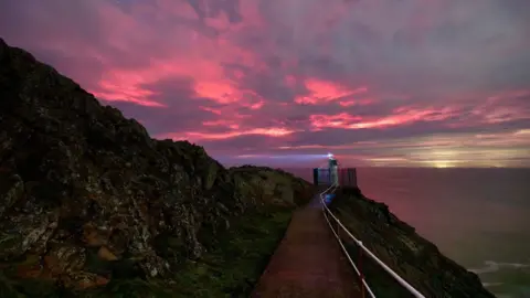 MATTHEW MUTTOCK Cloudy skies overt the edge of a cliff path. With red lights creeping through.