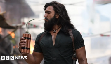 Ranveer Singh in a still from Dhurandhar, seen holding dynamite to light a cigarette. He's wearing a black shirt with a silver pendant around his neck. He has long hair and a beard.