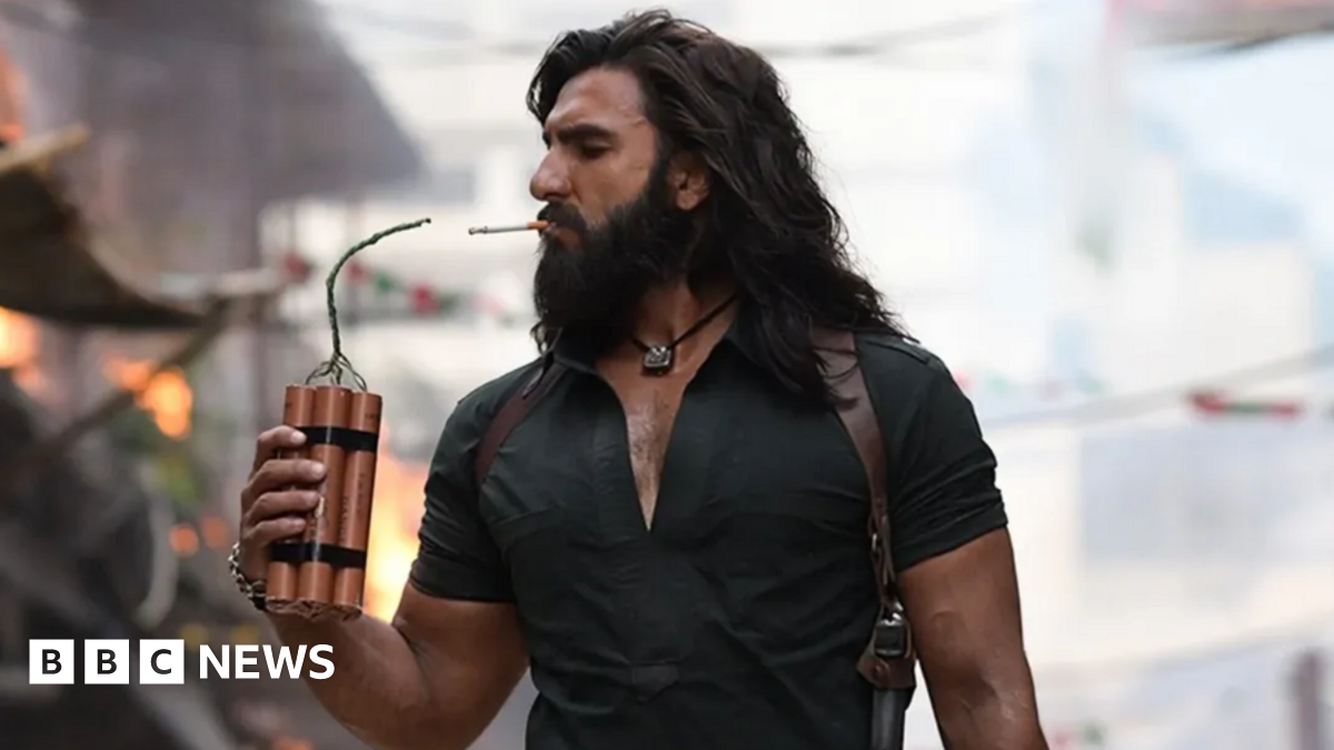 Ranveer Singh in a still from Dhurandhar, seen holding dynamite to light a cigarette. He's wearing a black shirt with a silver pendant around his neck. He has long hair and a beard.