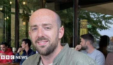 Oliver Long is looking at the camera. He is balding and has a beard and a moustache. He is wearing a green shirt with a white T-shirt underneath. Three men are sitting at tables in a cafe behind him.