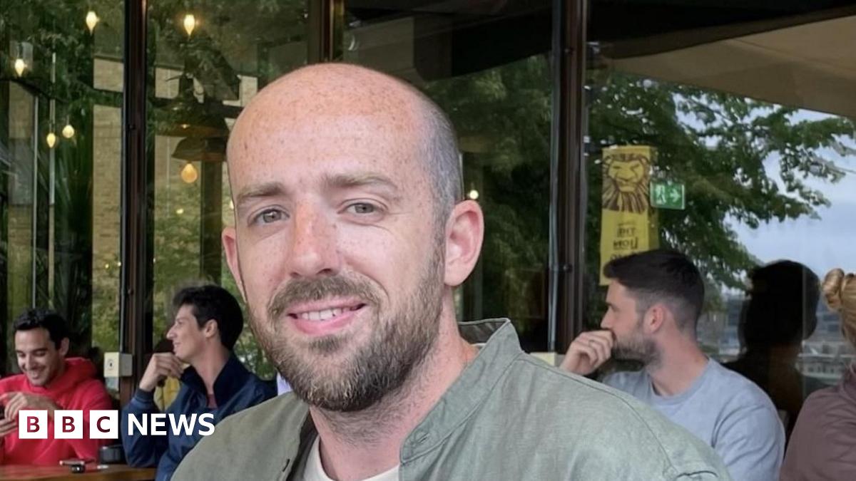 Oliver Long is looking at the camera. He is balding and has a beard and a moustache. He is wearing a green shirt with a white T-shirt underneath. Three men are sitting at tables in a cafe behind him.