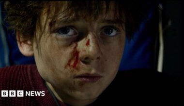 Alfie Williams is dressed as his character Spike from 28 Years Later: The Bone Temple. He looks directly into camera, and appears to be injured with blood on his face.