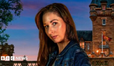 A close-up image of Rachel Duffy from County Down. She is looking at the camera with a neutral expression. She has long, dark hair and is wearing a blue denim jacket. Behind her is the traitors Castle, with the sky getting darker as the sun sets.