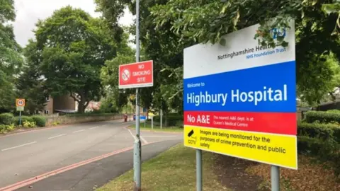 A sign by a roadside which reads HIGHBURY HOSPITAL NO A&E - THE NEAREST A&E DEPT. IS AT QUEEN'S MEDICAL CENTRE - IMAGES ARE BEING MONITORED FOR THE PURPOSES OF CRIME PREVENTION AND PUBLIC SAFETY. Another sign reads NO SMOKING SITE.