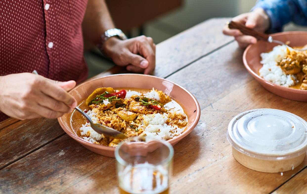 Man sitting at wooden table eating takeaway curry with rice, container next to plate