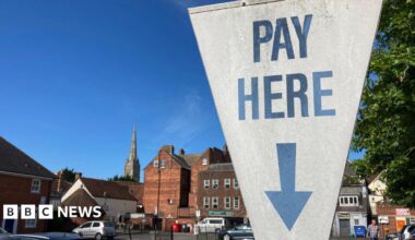 A white sign at a car park in Salisbury, Wiltshire, which reads "pay here" in blue writing, with a blue arrow pointing down. The sign is shaped like an upside down triangle. Cars can be seen parked in the city car park behind, which is surrounded by older buildings and a large spire can be seen in the background. It is a sunny day.