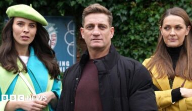 Jennifer Metcalfe, who is wearing a grey tracksuit and red top, is standing next to Nikki Shadwick, who is wearing a cream jumper, with her arm around her shoulder on the set of Hollyoaks,