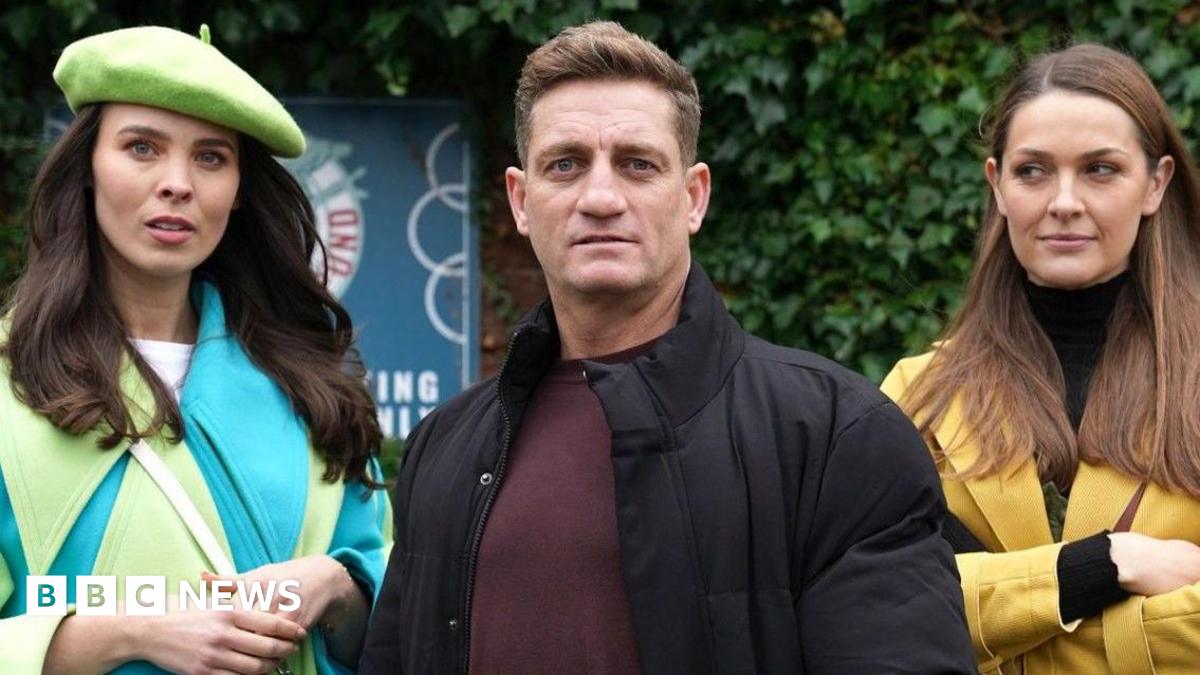 Jennifer Metcalfe, who is wearing a grey tracksuit and red top, is standing next to Nikki Shadwick, who is wearing a cream jumper, with her arm around her shoulder on the set of Hollyoaks,