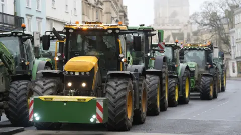 PA Media A queue of green and yellow tractors makes its way through an Oxford city centre road, with historic buildings in the background, under a misty sky